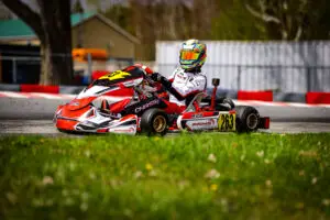 Go-kart racer in a white suit with a colorful, mirrored helmet driving a red-and-white kart on an outdoor track.