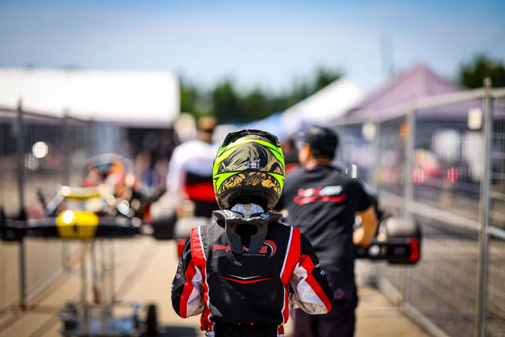 Back of a rider wearing a neon yellow helmet and red-black racing suit in the pit lane, with blurred pit area in the background.