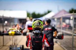 Back of a rider wearing a neon yellow helmet and red-black racing suit in the pit lane, with blurred pit area in the background.