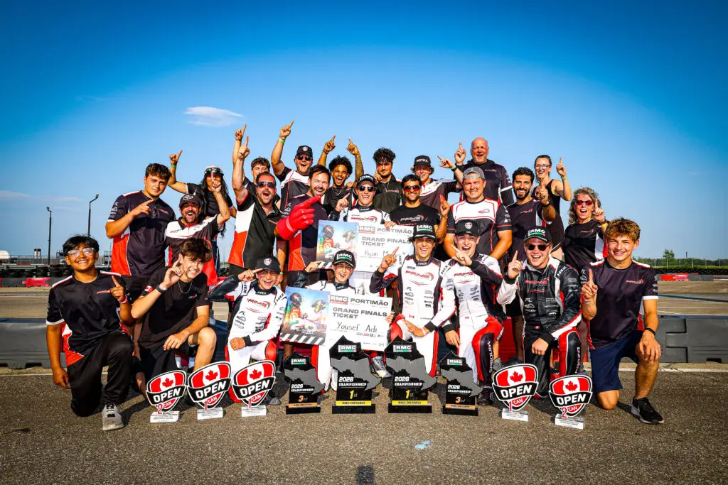 Group of racers posing together on a track, raising one finger in celebration with trophies and banners under a bright blue sky.