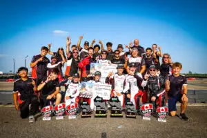 TEAM PRIME-26-CAN-OPEN-CKN-13967 - Prime Powerteam - Prime Powerteam Group of racers posing together on a track, raising one finger in celebration with trophies and banners under a bright blue sky.