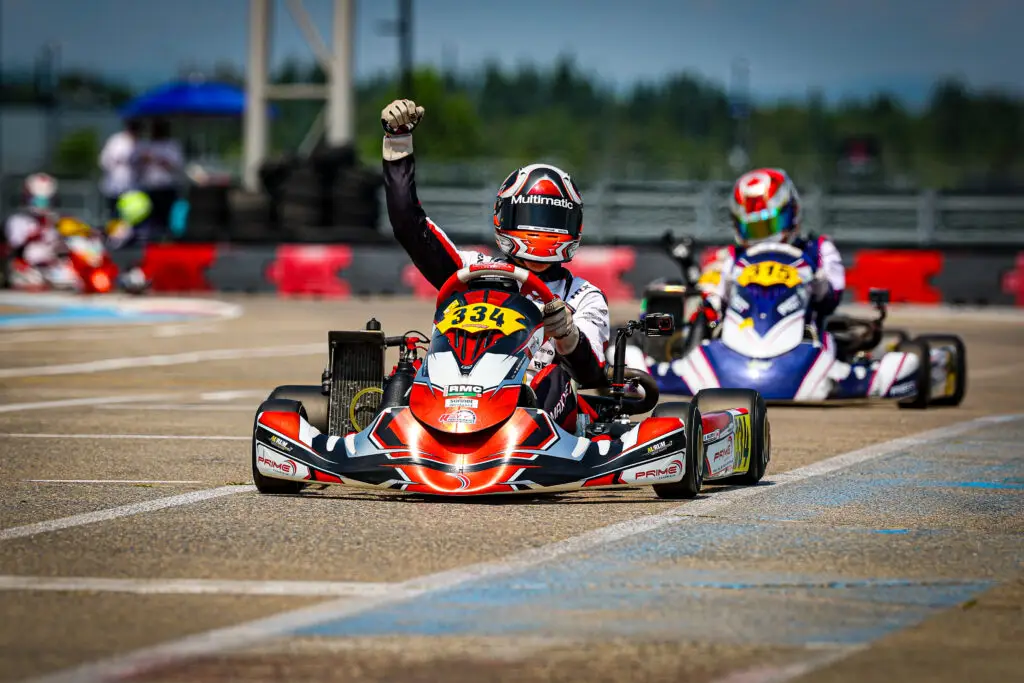 Go-kart racer in red-and-white suit raises a fist while driving on a track; number 334 on the front kart with other racers blurred in the background.