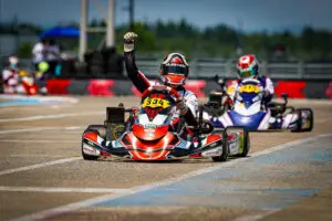 26-CAN-OPEN-CKN-12008 - Prime Powerteam - Prime Powerteam Go-kart racer in red-and-white suit raises a fist while driving on a track; number 334 on the front kart with other racers blurred in the background.